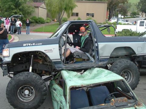 1989 Ford Bronco - A lifted truck is shown crushing a smaller vehicle, with onlookers in the background.