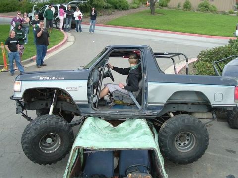 1989 Ford Bronco - A heavily modified Ford Bronco with oversized tires is shown driving over a smaller vehicle.