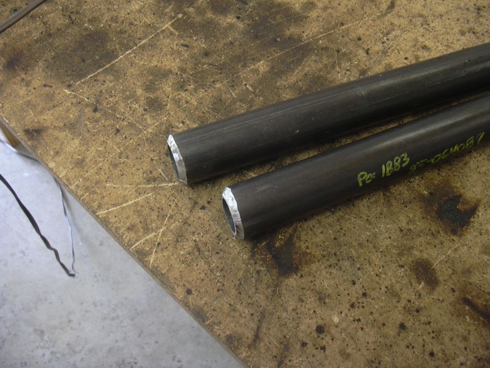 Two metal tubes with markings 'Po: 1883' and '37-054087' on a workbench