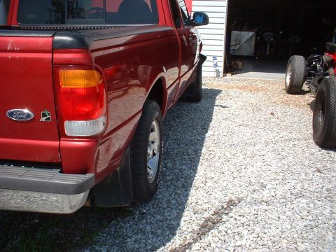 1999 Ford Ranger - A red Ford Ranger pickup truck is seen from the rear quarter angle, parked on a gravel surface outside a garage.