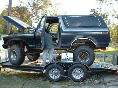 1978 Ford Bronco - A black 1978 Ford Bronco is on a trailer with its hood open and driver's door ajar as someone works on it.