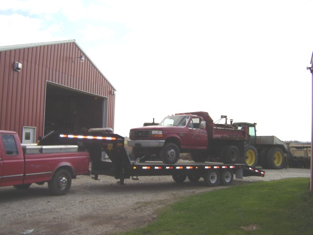 Red dump truck loaded on a trailer, with a tractor behind it. A red pickup truck is towing the trailer