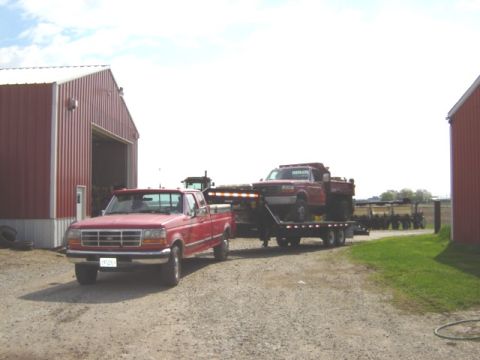 1992 Ford Bronco - A red Ford pickup truck tows a trailer carrying another truck, with farm buildings in the background.