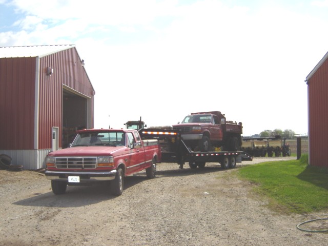 Red Ford F-250 pickup truck towing a flatbed trailer with a red Ford F-350 dump truck on it, parked in front of a barn