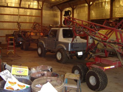 1992 Ford Bronco - A Suzuki Samurai is parked inside a barn, connected to a large red sprayer.