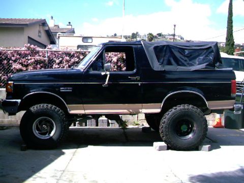 1989 Ford Bronco - This black Ford Bronco has a tan stripe and a black soft top, seen from the side.