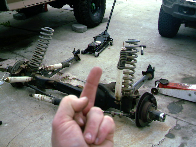 vehicle suspension parts on ground, jack nearby, another vehicle in background