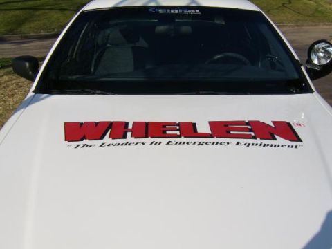 1999 Ford Crown Victoria - A white car hood displays prominent red lettering for "WHELEN" and a smaller tagline about emergency equipment.