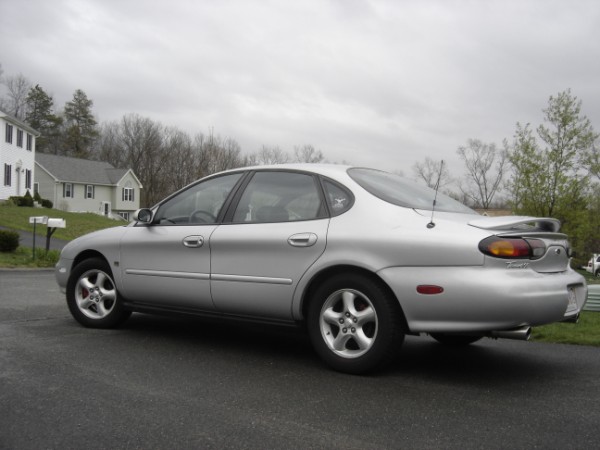 Silver 1999 Ford Taurus sedan, rear 3/4 view, parked on asphalt with houses and trees in background