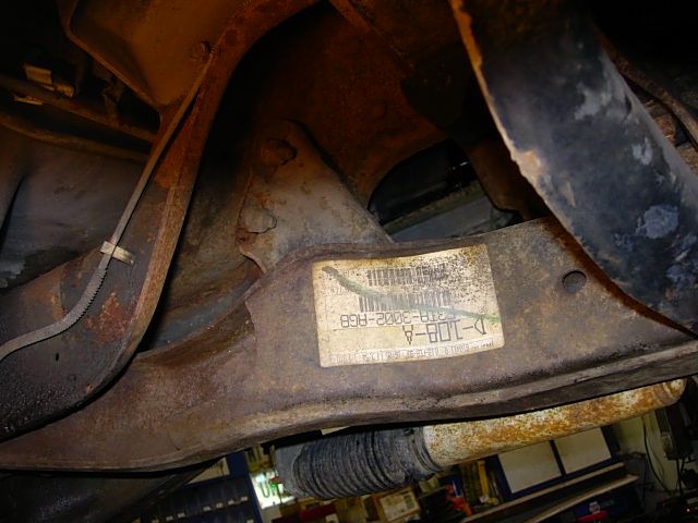 Underside of a vehicle showing rusty suspension parts and a sticker with part numbers like D-108-A and 37B-3002-RG