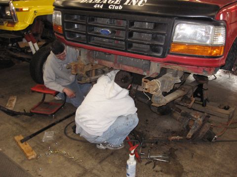 1977 Ford F-150 - Two mechanics work on the front suspension of a red Ford truck, with a yellow Jeep visible in the background.