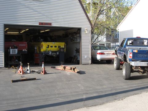1977 Ford F-150 - A lifted blue pickup truck with prominent tires is parked in front of a garage, with other vehicles visible inside and nearby.