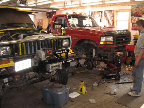 1977 Ford F-150 - Two trucks, a yellow Jeep and a red Ford, are in a garage with parts removed and tools scattered around them.