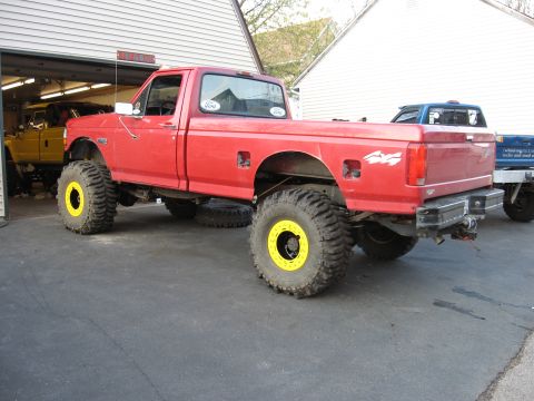 1977 Ford F-150 - A lifted red pickup truck with oversized tires and yellow wheel accents sits in a driveway.