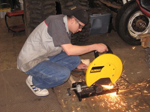 1977 Ford F-150 - A person is using a chop saw to cut metal in a workshop environment with large tires visible.