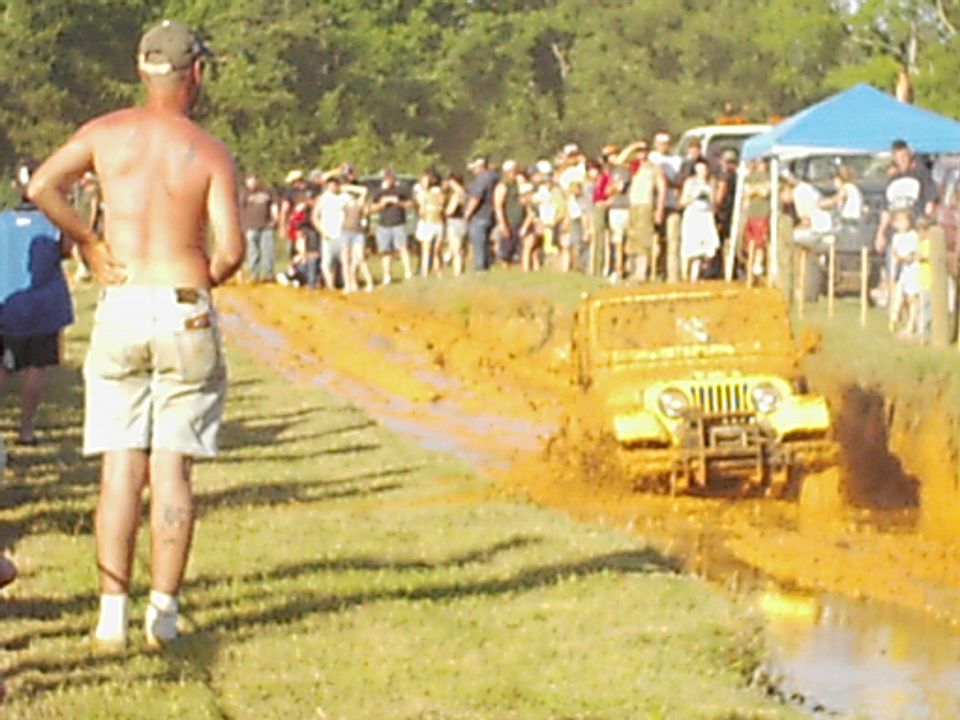 Yellow jeep struggling through a mud pit with spectators watching
