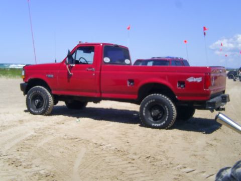 1977 Ford F-150 - A red pickup truck is parked on a sandy area near the beach, with another vehicle visible behind it.