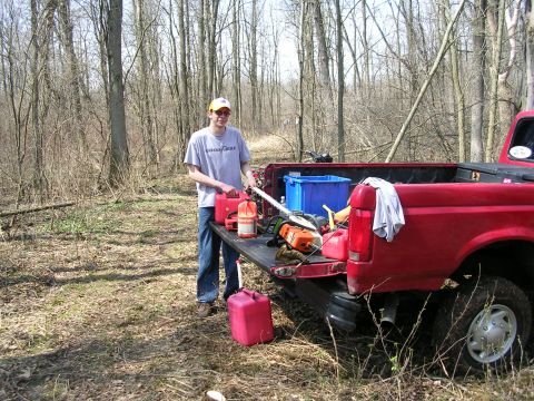 1977 Ford F-150 - Person with tools and gas cans next to the bed of a red pickup truck in a wooded area.