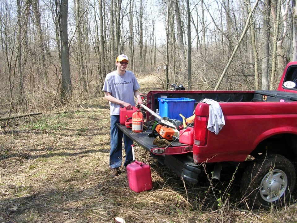 Person standing by the open tailgate of a red pickup truck, holding a red gas can. Chainsaws and a blue bin are in the truck bed
