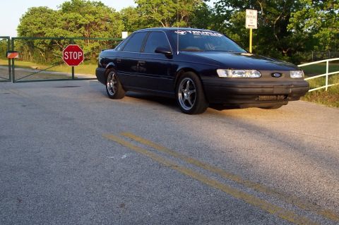 1993 Ford Taurus - A dark blue 1993 Ford Taurus is parked near a stop sign and gate, with a "TAURUS" banner on its windshield.