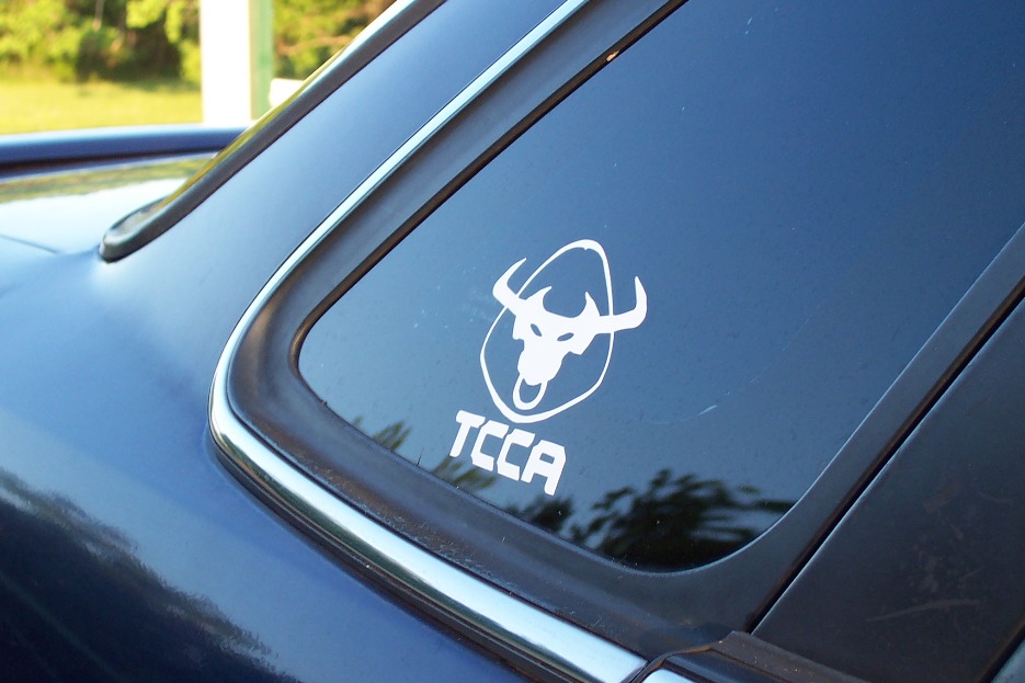 Blue car rear window with white TCCA bull logo sticker. Supermotors.net watermark at bottom right