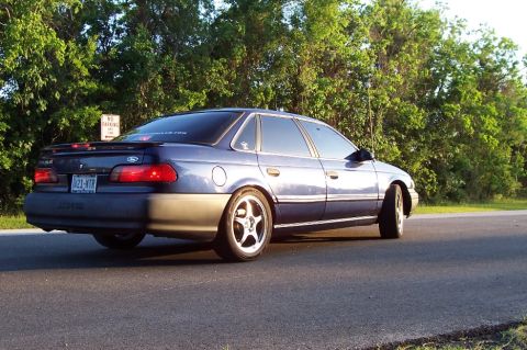 1993 Ford Taurus - Rear view of a blue sedan with aftermarket wheels, parked on a road with trees in the background.