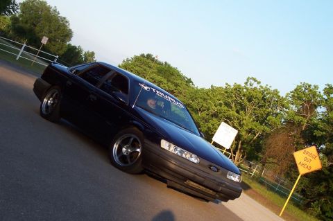 1993 Ford Taurus - A dark blue sedan with aftermarket wheels is parked on the side of a road with trees in the background.