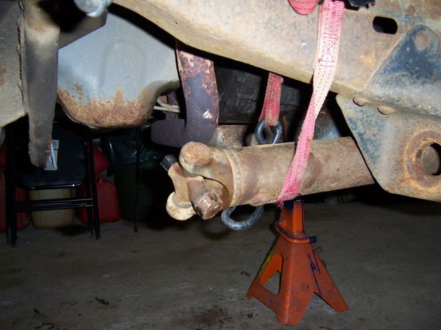 Rusty solid front axle on orange jack stand with pink strap, underbody garage shot