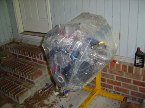 1986 Ford Bronco - Engine on a stand, wrapped in plastic, sits on a porch next to brick steps.