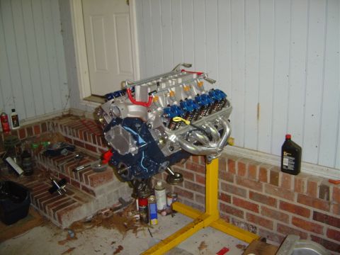 1986 Ford Bronco - A V8 engine with chrome headers and blue valve covers is mounted on a yellow engine stand.