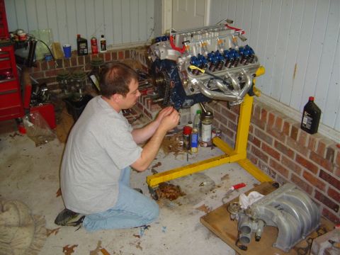1986 Ford Bronco - A man works on a V8 engine mounted on a yellow stand in a garage. The engine appears to be a Ford V8.