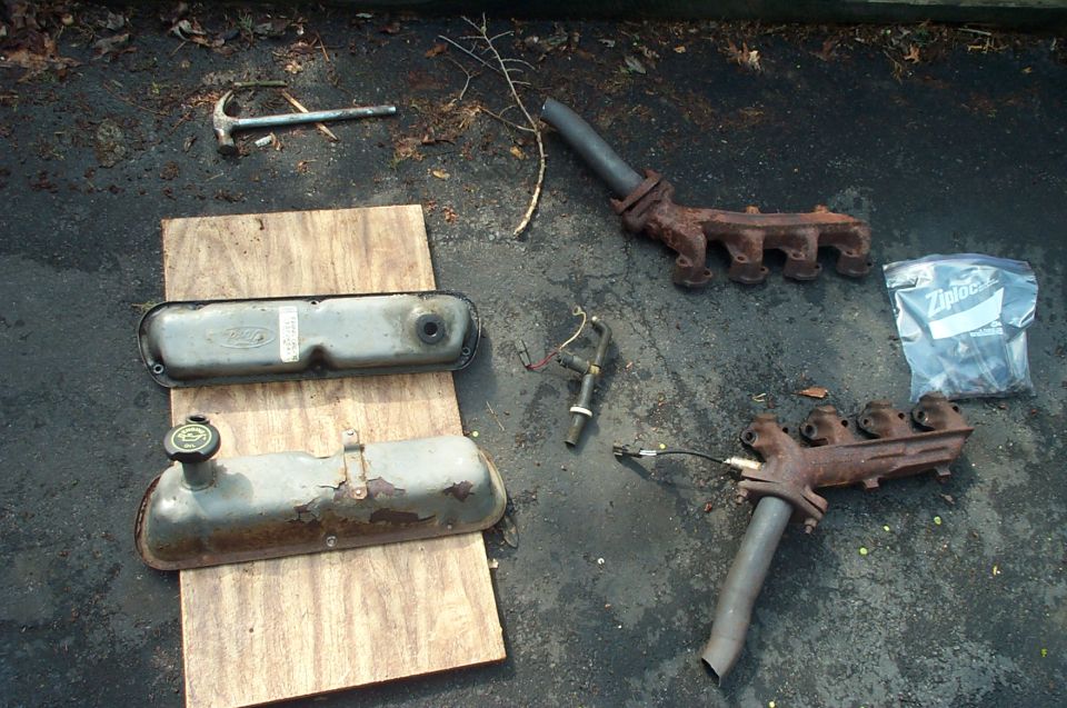 Ford valve covers, rusty exhaust manifolds, and hardware laid out on pavement.