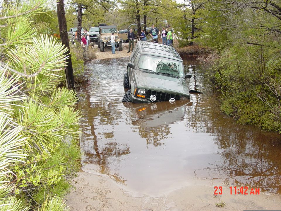 Green Jeep Cherokee partially submerged in muddy water, front grille and headlights visible