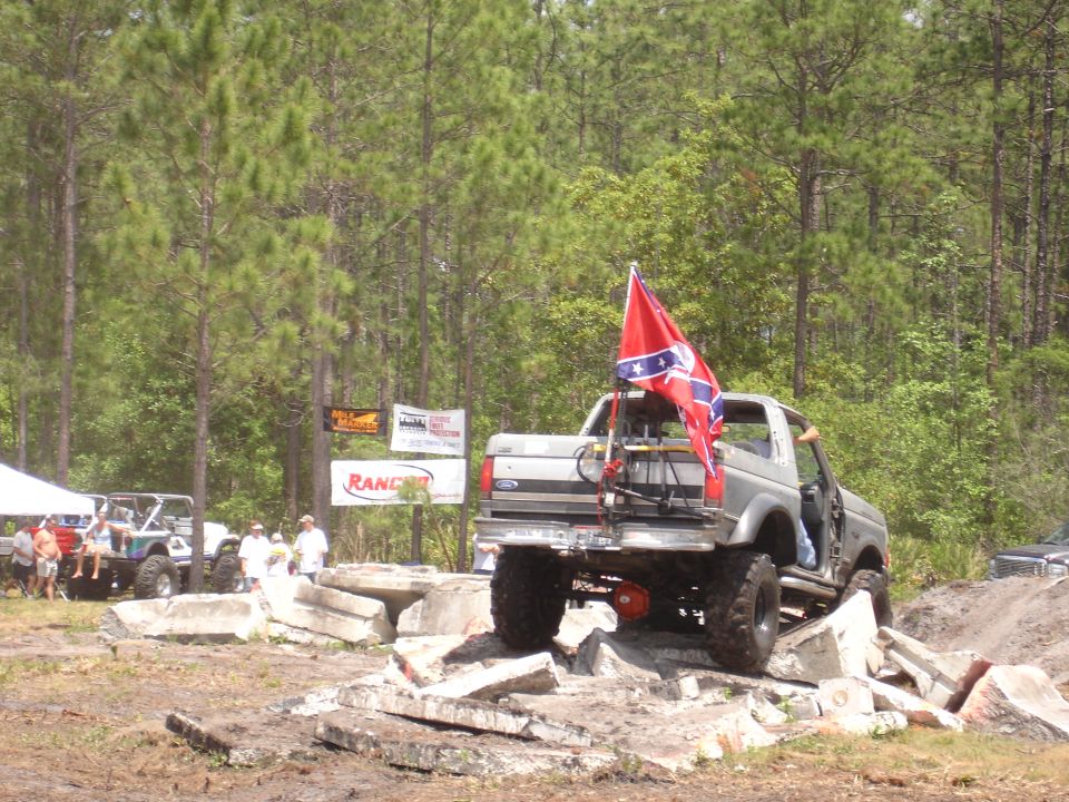 Silver pickup truck with a Confederate flag on the back, driving over concrete obstacles. Another off-road vehicle is visible to the left