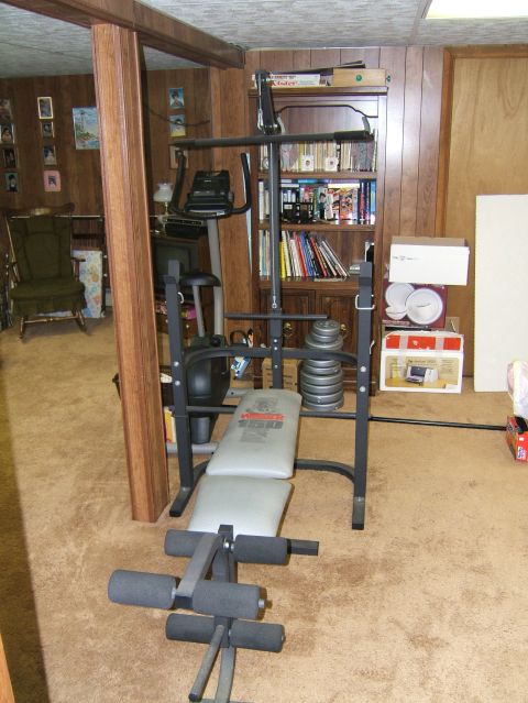 1998 Ford Taurus - A home gym setup a weight bench and stationary bike is shown in a basement room with wood paneling and carpet.
