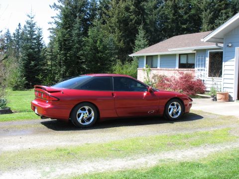 1997 Ford Probe - Red Ford Probe GT hatchback coupe with rear spoiler and chrome wheels parked on a gravel driveway by a blue house.