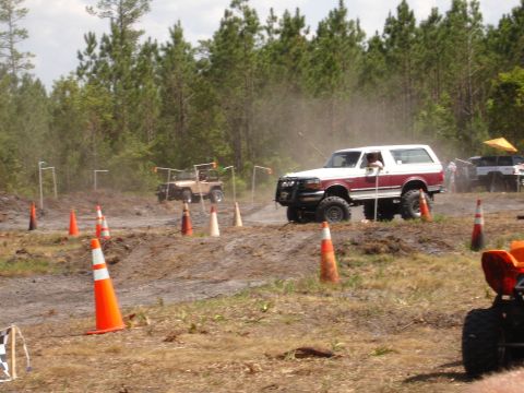 1989 Ford Bronco - Two off-road vehicles drive through a muddy course marked with cones.