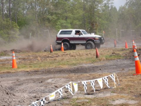 1989 Ford Bronco - A two-tone maroon and white Ford Bronco navigates a dusty off-road course marked by orange cones.