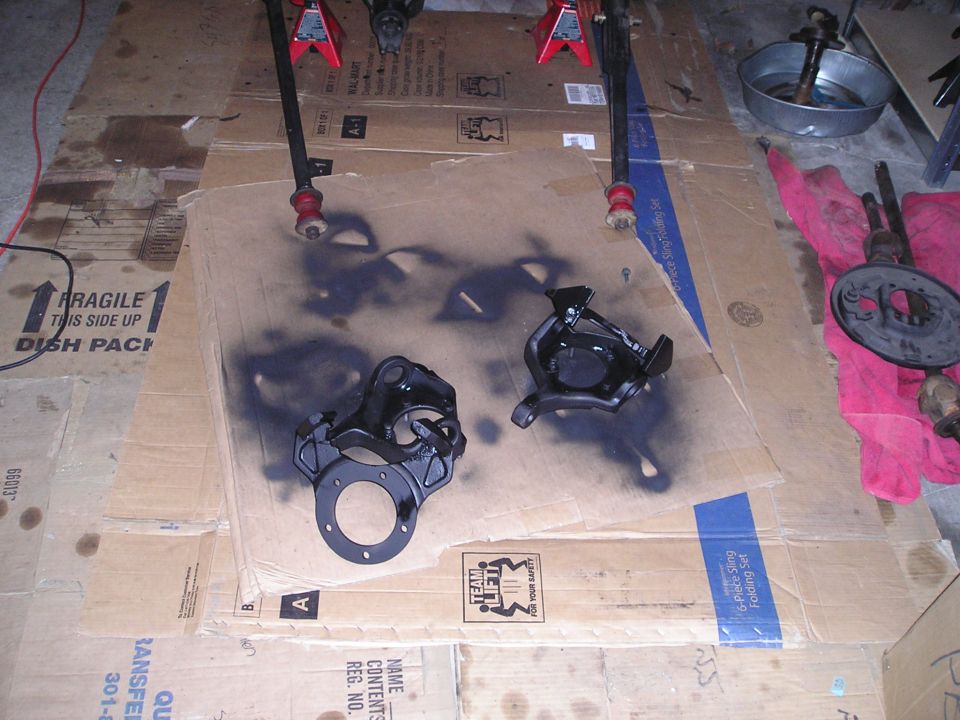Two black painted car parts on cardboard with spray paint overspray, jack stands, and a brake rotor