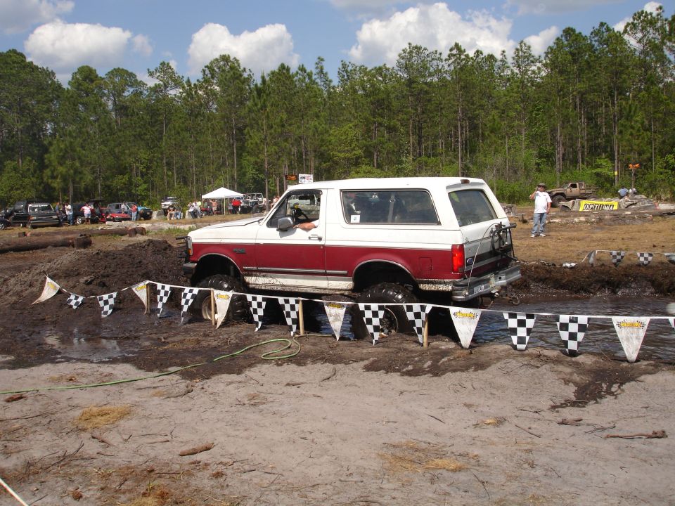 Two-tone red and white Ford Bronco going through a mud pit with checkered flags