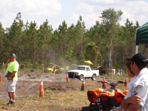 1989 Ford Bronco - A white pickup truck jumps over a muddy obstacle during an off-road event.