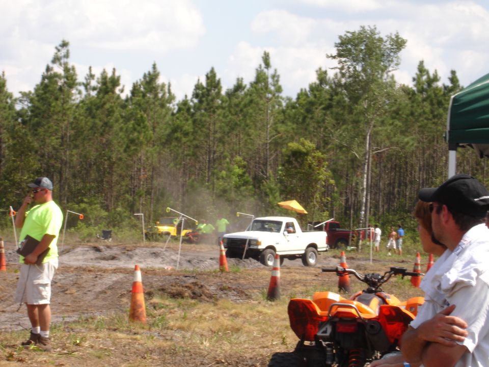 White pickup truck with a yellow canopy on its bed, mid-air over a muddy track. Its front grille is visible