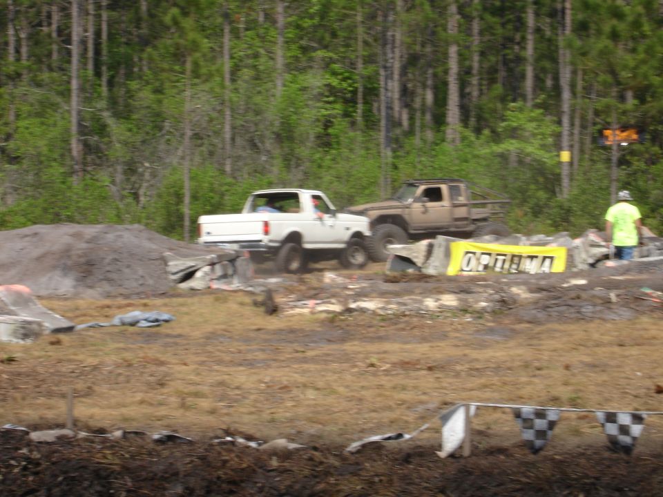 White pickup truck and tan off-road truck in muddy terrain with OPTIMA banner