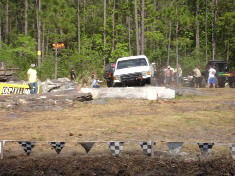 1989 Ford Bronco - A white pickup truck attempts to clear an obstacle during an off-road event, with other vehicles and spectators present.