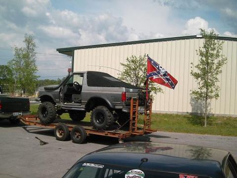 1986 Ford Bronco - A modified Ford Bronco is loaded onto a trailer, parked in front of a building with a Confederate flag flying nearby.