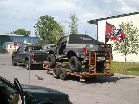 1986 Ford Bronco - A lifted 1986 Ford Bronco is seen on a trailer, towed by a pickup truck, with a Confederate flag flying.