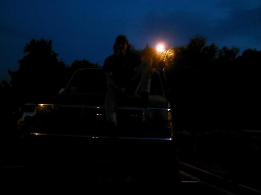 Person sits on the hood of a dark-colored truck at night, illuminated by a distant light