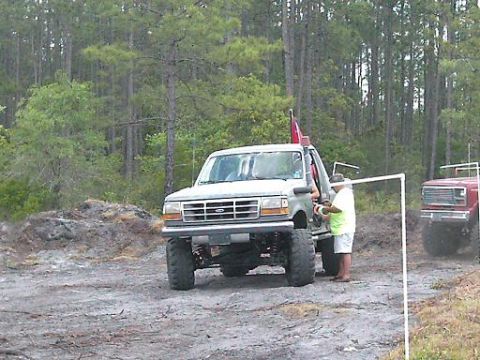 1986 Ford Bronco - A lifted light-colored truck with a red flag is parked next to a red truck in a wooded area.