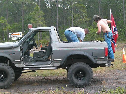 Gray Ford Bronco with off-road tires and a snorkel, doors removed, two men working on it, Confederate flag flying