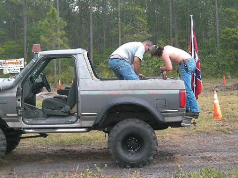 1986 Ford Bronco - Two men work on a gray 1986 Ford Bronco, which is parked in a muddy, wooded area with a Confederate flag flying nearby.